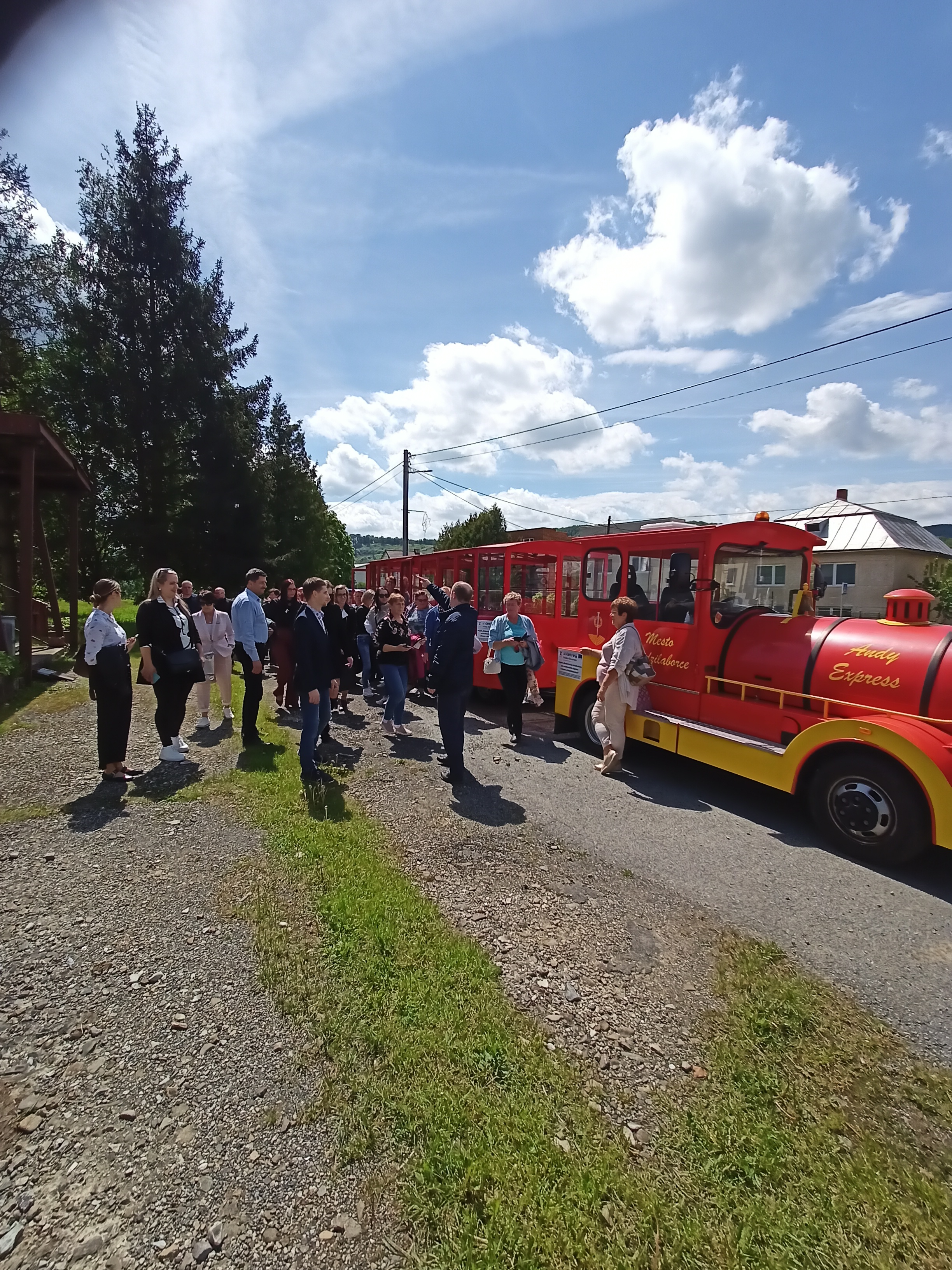 Grupa ludzi wsiada do czerwono-ż&oacute;łtego pociągu turystycznego "Andy Express". Słoneczny dzień, drzewa i budynki w tle.